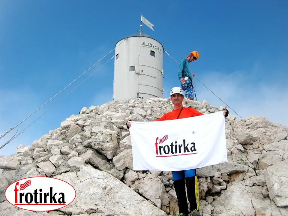 Алпинист на врвот Триглав 2864 м.н.в. во Јулиски Алпи држи бел пешкир со логото на Фротирка како дел од експедицијата Трилогија Алпи.