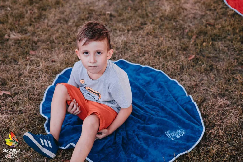 Дете седи на сина кружна Jolly фротирска прекривка со вез Фротирка & SOF за време на Струмица Опен Фест 2020 во градскиот парк.