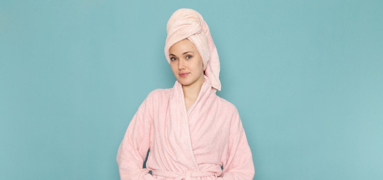 Жена во мека розова фротирна бањарка и пешкир на глава, симбол на удобност, елеганција и топлина.