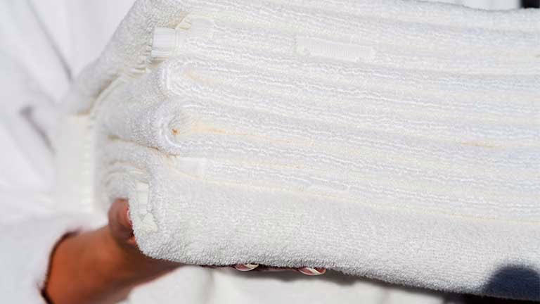 Сет од бели пешкири за бања држени во рака, симбол на свежина и мекост, со совети за враќање на нивната апсорбентност и мекост.