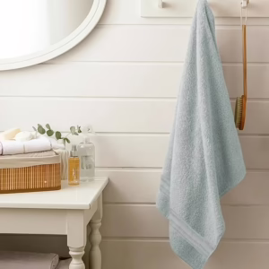 Светлосин пешкир за бања закачен на ѕидна закачалка покрај масичка со козметика и огледало во модерна бања.